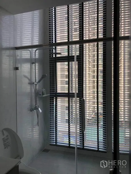 Condominium for sale in Ratchathewi, Bangkok - Bathroom with glass shower enclosure, handheld shower and tall windows with blinds.