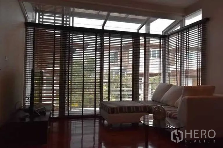 Detached House for sale in Bueng Kum, Bangkok - Sunroom lounge with L-shaped sofa, floor-to-ceiling glass and venetian blinds overlooking the community.