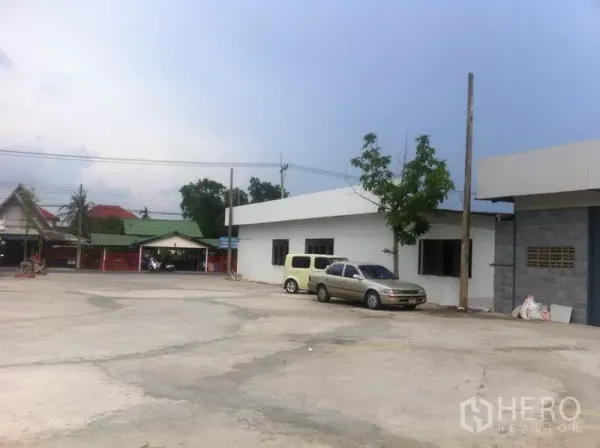 Warehouse for rent in Lam Luk Ka, Pathum Thani - Spacious concrete yard with parking beside a low-rise warehouse and nearby neighborhood houses in Lam Luk Ka.
