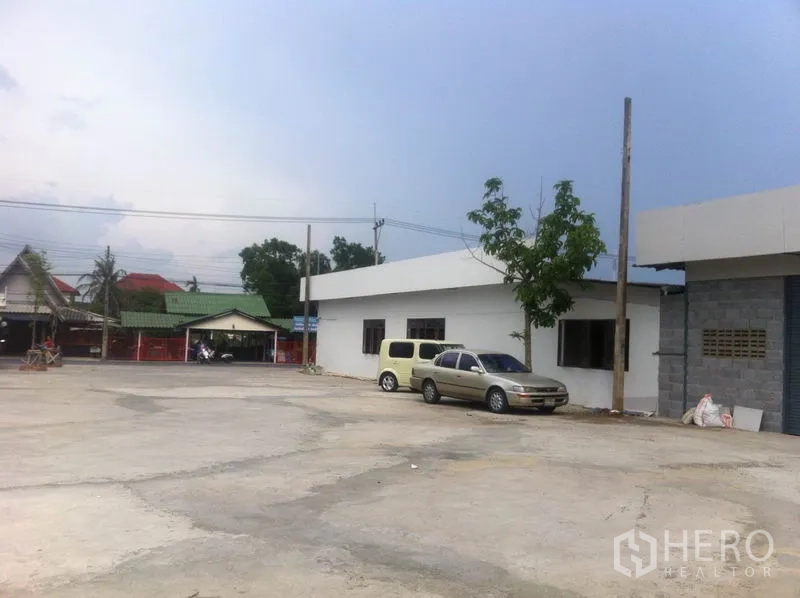 Warehouse for rent in Lam Luk Ka, Pathum Thani - Spacious concrete yard with parking beside a low-rise warehouse and nearby neighborhood houses in Lam Luk Ka.