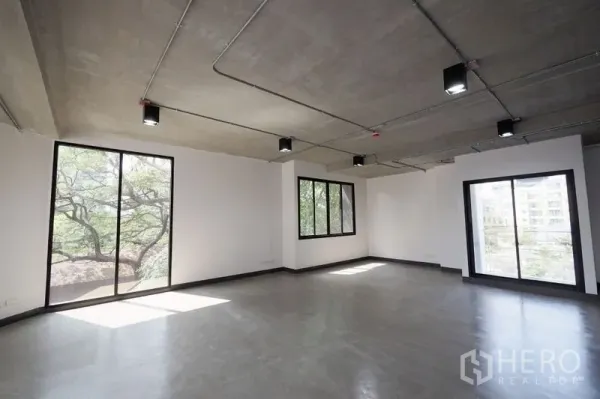 Office Space for rent in Watthana, Bangkok - Open-plan office with polished concrete floors, exposed ceiling lights, and large windows with tree views.