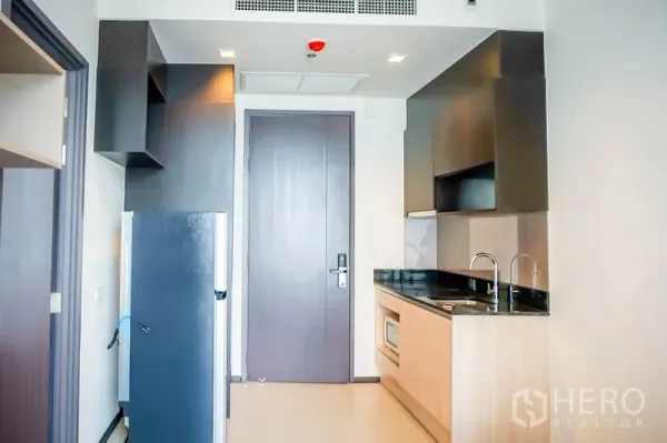 Condominium for sale in Watthana, Bangkok - Compact kitchenette with black countertop, sink, microwave and refrigerator by the entry.