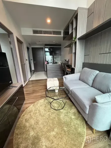 Condominium for rent or sale in Watthana, Bangkok - Sofa corner with built-in shelving, round rug and TV unit by a large window.