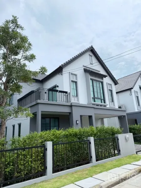 บ้าน ให้เช่าในLat Krabang, Bangkok - ภาพมุมเอียงของตัวบ้าน มีระเบียง รั้วเหล็กสีดำ และแนวต้นไม้พุ่มจัดสวน