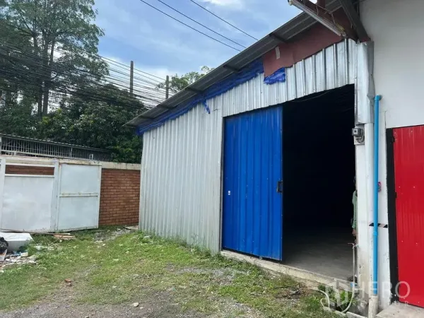 คลังสินค้า ให้เช่าในMueang Samut Prakan, Samut Prakan - ภายนอกโกดังผนังเมทัลชีท ประตูบานเลื่อนสีน้ำเงิน ข้างกำแพงอิฐและต้นไม้
