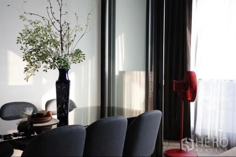 คอนโด ให้เช่า - มุมทานอาหารรับแสงพร้อมแจกันสีน้ำเงิน เก้าอี้สีเทา และพัดลมสีแดง