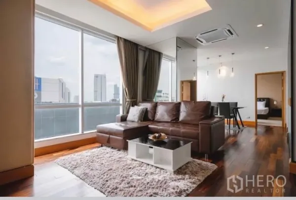 Condominium for rent in Sathon, Bangkok - Bright living room with floor-to-ceiling windows, brown leather sofa, recessed ceiling lights, hardwood floors and city skyline view.
