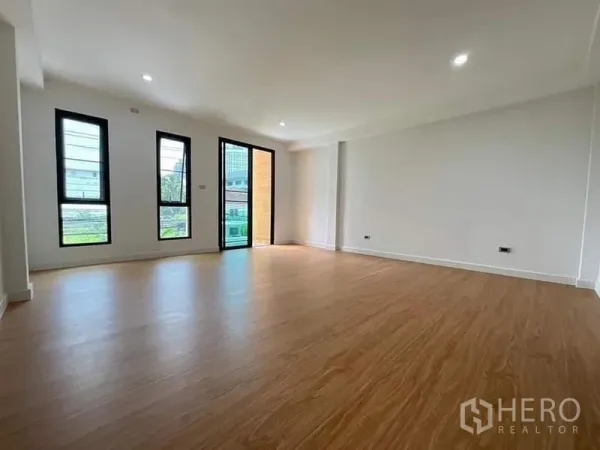 Townhouse for sale in Watthana, Bangkok - Bright living area with wood-look flooring, recessed lighting and tall windows opening to a balcony.