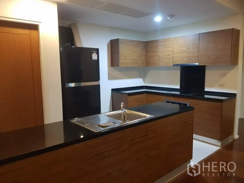 Condominium for rent in Watthana, Bangkok - Modern L-shaped kitchen with wooden cabinets, black stone countertops, island sink, and a full-size refrigerator.