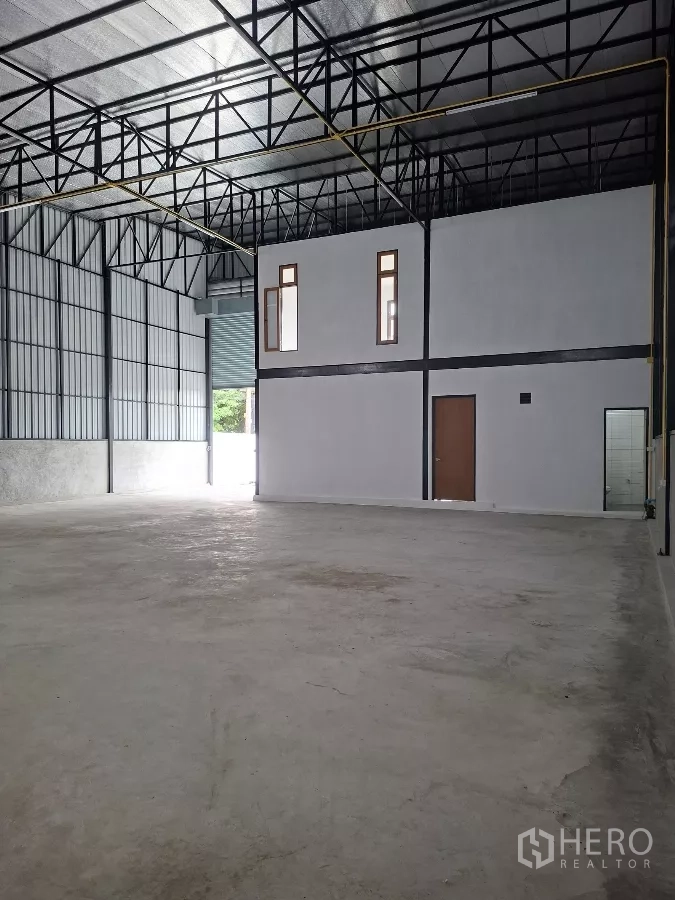 Warehouse for rent in Lam Luk Ka, Pathum Thani - Open warehouse bay with high steel truss roof, partitioned two-storey office block and concrete floor near the loading door.