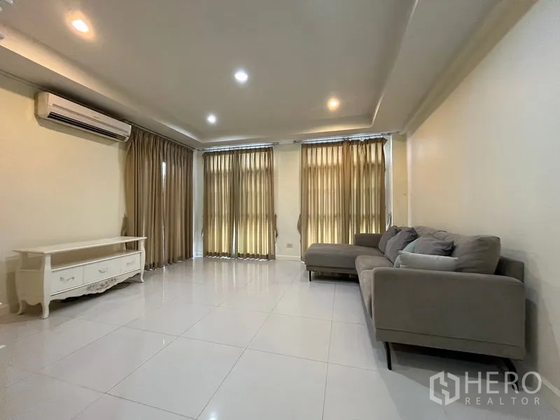 Townhouse for rent in Phaya Thai, Bangkok - Spacious living room with L-shaped sofa, recessed ceiling lights, air conditioner and tall curtains over glossy tiles.