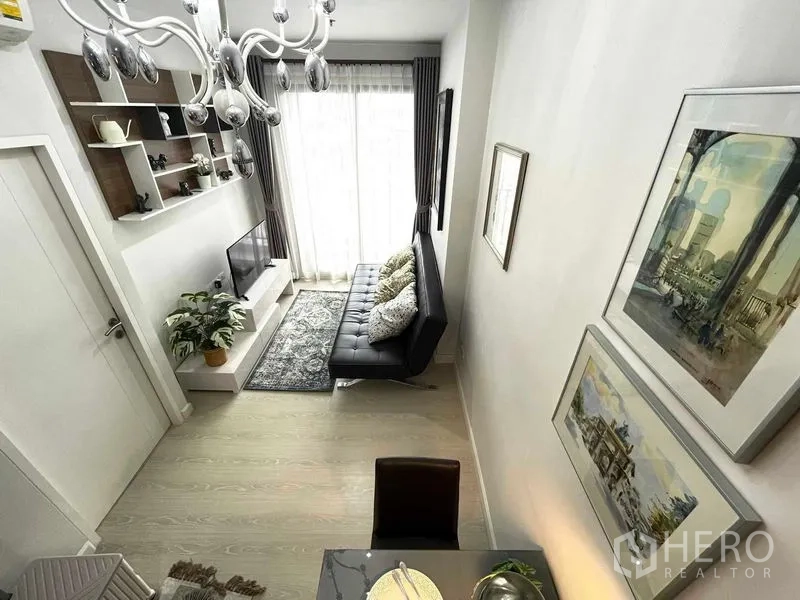Condominium for sale in Huai Khwang, Bangkok - Light-filled living room with black sofa, TV console, wall shelves and sheer curtains to the balcony.