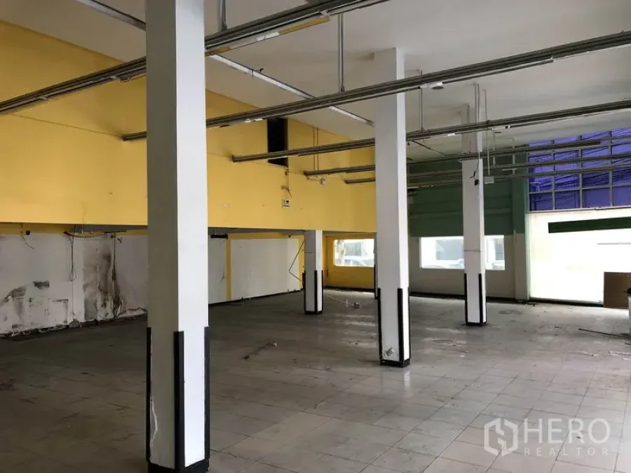 Warehouse for rent in Suan Luang, Bangkok - Open-plan warehouse interior with white columns, yellow accent walls, tiled flooring, and large front windows allowing natural light.