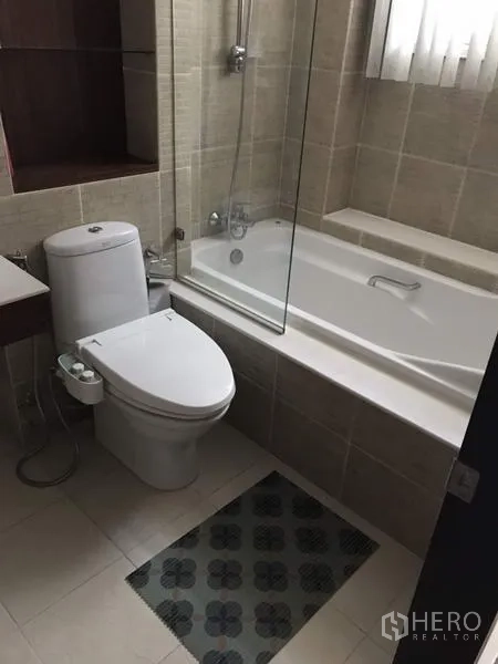 Condominium for rent in Watthana, Bangkok - Bathroom with bathtub and glass shower screen, handheld shower, toilet and beige wall tiles.