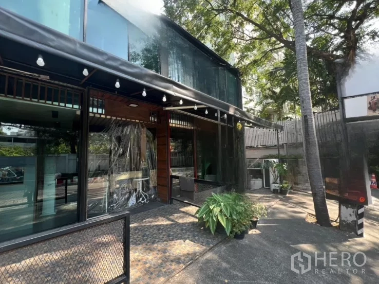 零售空间 出租在Watthana, Bangkok - 玻璃外立面侧视图，带遮荫露台、灯串、树木以及场内停车位。