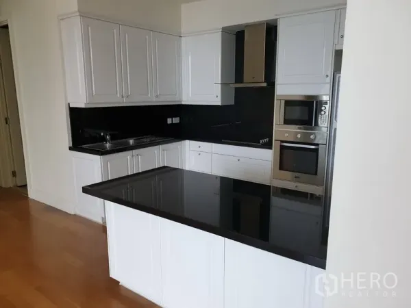 Condominium for rent in Watthana, Bangkok - Modern L-shaped kitchen with white cabinetry, black stone countertop, built-in oven, microwave, and cooktop.