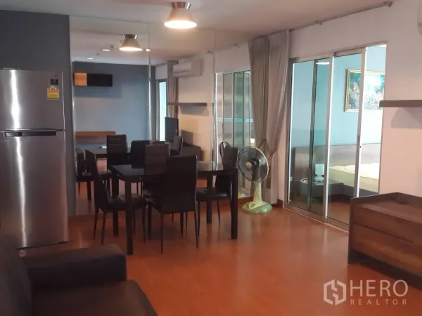 Condominium for rent in Huai Khwang, Bangkok - Open-plan living and dining space with stainless refrigerator, black dining set, wood floors and glass doors leading to the bedroom.