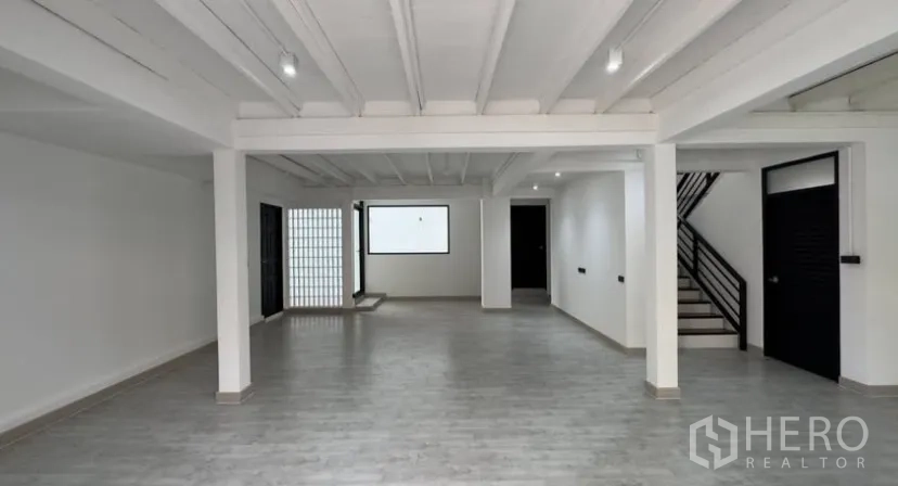 Detached House for rent in Suan Luang, Bangkok - Open-plan living area with grey wood floors, white beamed ceiling, black doors and a side staircase.