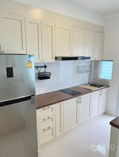 联排别墅 出租在Prawet, Bangkok - 一字型厨房，白色橱柜、木质台面、瓷砖背墙、灶台与不锈钢冰箱。