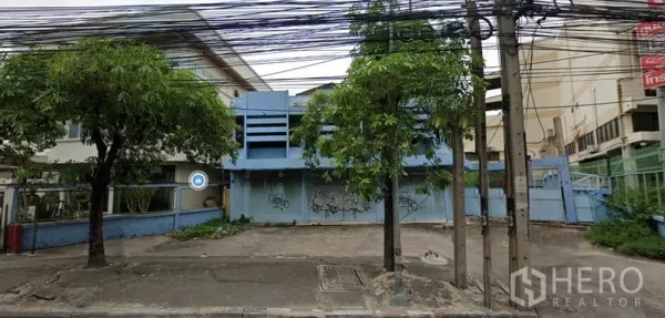 Shophouse for rent in Bang Phli, Samut Prakan - Street view of a blue two-storey shophouse in Bang Phli with roller shutters, a paved forecourt for parking, roadside trees, and overhead utility lines.