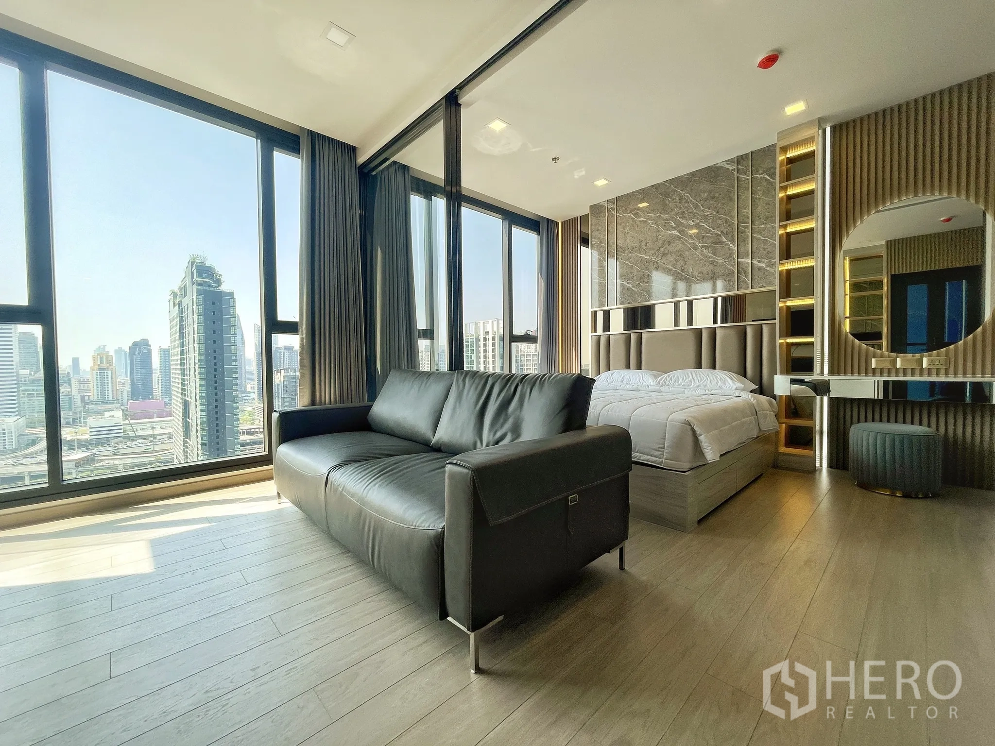 公寓 for rent in Huai Khwang, Bangkok - 明亮的公寓起居与卧室一体空间，黑色沙发、床、落地窗与城市天际线景观，内置梳妆台与木地板。