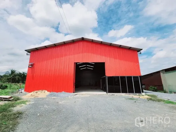 Warehouse for rent in Sam Phran, Nakhon Pathom - Approach to the red warehouse showing the open bay and gravel driveway under a cloudy sky.