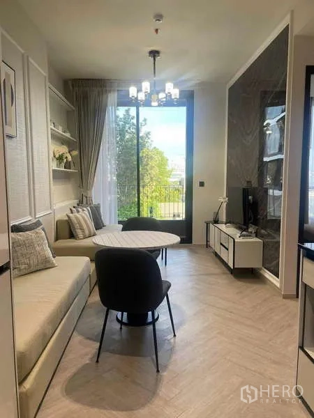 คอนโด ให้เช่า - ห้องนั่งเล่นเปิดโล่ง โคมระย้า โซฟาบิวท์อิน ตู้ทีวี และประตูกระจกออกระเบียงเห็นต้นไม้