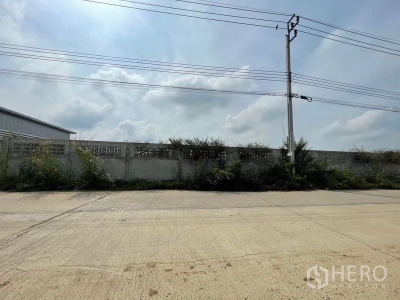 Land for sale in Sai Noi, Nonthaburi - Frontage of the land facing a paved road with electricity poles and boundary wall.
