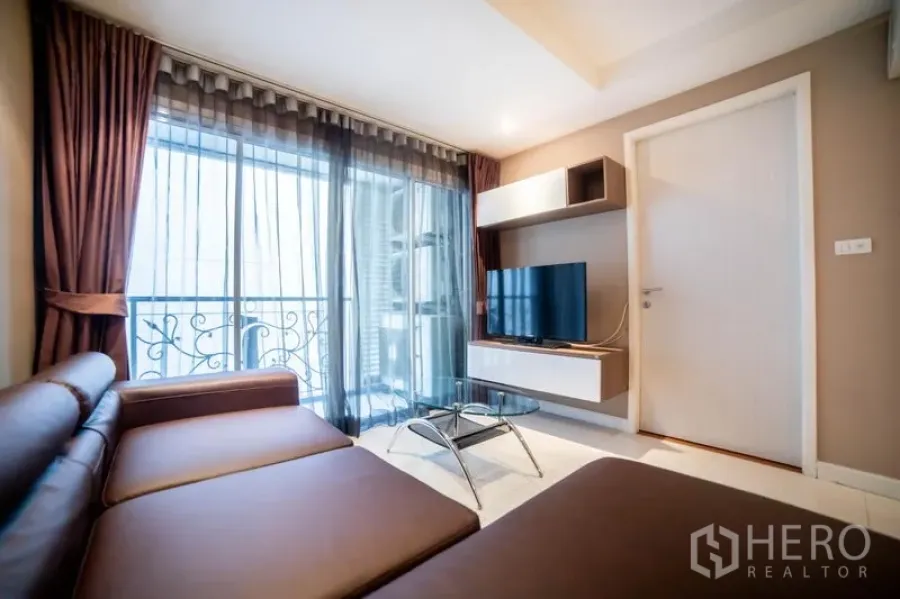 คอนโด ให้เช่าในWatthana, Bangkok - ห้องนั่งเล่นอบอุ่น โซฟาเข้ามุม ทีวีชั้นลอย โต๊ะกลางกระจก และระเบียงหลังผ้าม่านโปร่ง