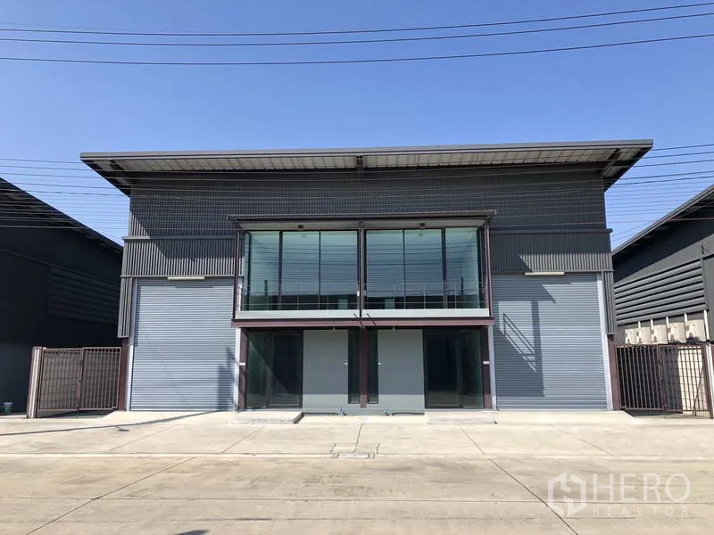 货栈 出租在Bang Phli, Samut Prakan - 现代灰色仓库立面，玻璃办公室、双卷闸门和宽阔混凝土前院。