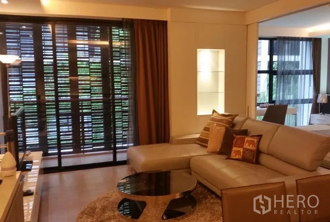 Condominium for rent or sale in Watthana, Bangkok - Lounge area with sectional sofa, round glass coffee table and balcony screened for privacy; bedroom beyond.