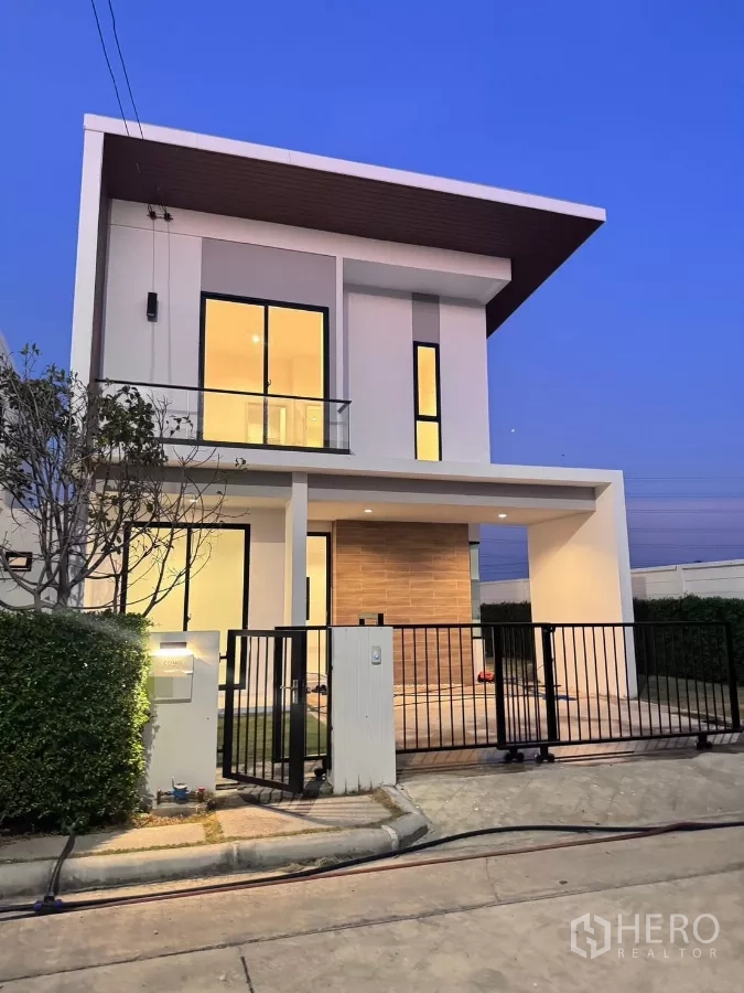 Detached House for rent in Samut Prakan - Modern two-storey detached house with slanted roof, warm lights and black gate at dusk.