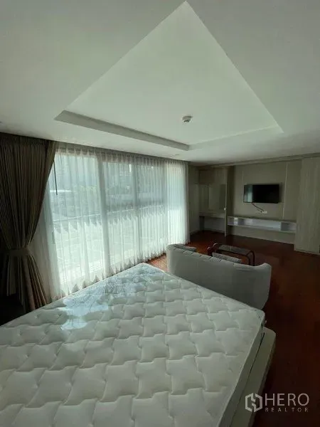 Condominium for rent in Watthana, Bangkok - Spacious master bedroom with sofa seating, TV wall unit and wide windows with sheer curtains and wood floors.