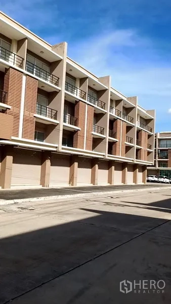 Office Space for sale in Khlong Luang, Pathum Thani - Row of modern four‑storey commercial offices with balconies and roller shutters along a wide driveway under a blue sky.