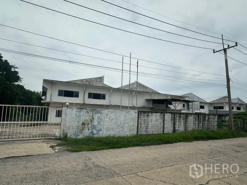 Factory for rent or sale in Khlong Luang, Pathum Thani - Streetside view of a gated standalone factory complex with perimeter wall and power lines in Khlong Luang.