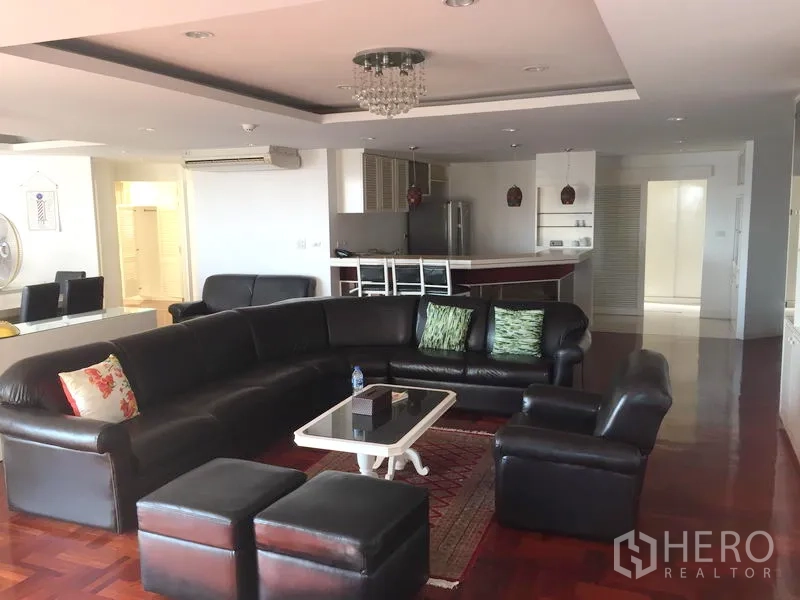 Condominium for rent in Watthana, Bangkok - Spacious open-plan living room with black leather sectional, chandelier, and hardwood floors facing the kitchen.