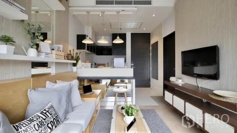 Condominium for rent or sale in Khlong Toei, Bangkok - Bright open-plan living room with built-in shelves, TV wall, and breakfast bar leading to the kitchen.