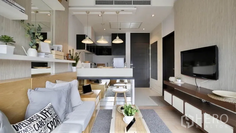 Condominium for rent or sale in Khlong Toei, Bangkok - Bright open-plan living room with built-in shelves, TV wall, and breakfast bar leading to the kitchen.