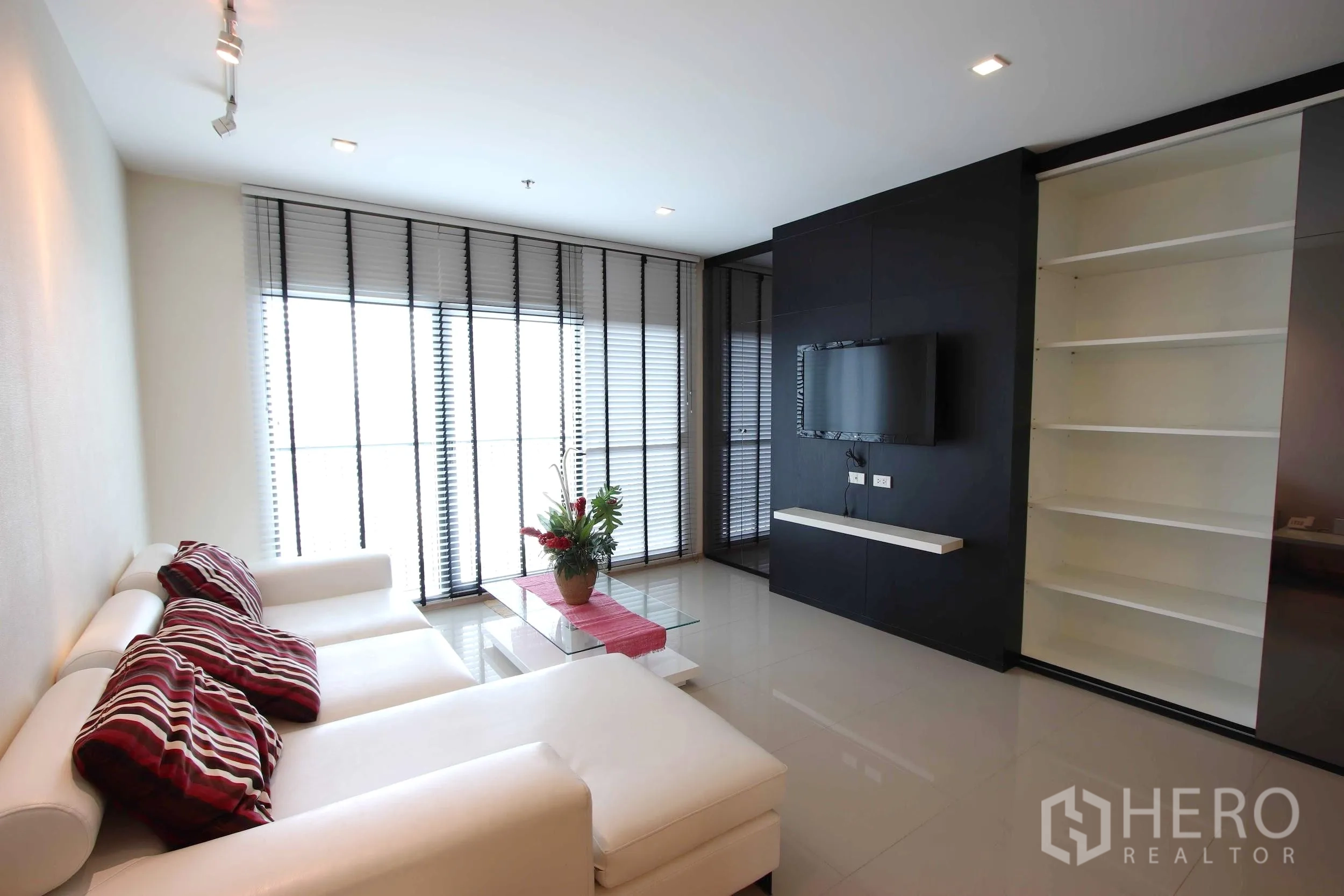 คอนโด ให้เช่าในKhlong Toei, Bangkok - ห้องนั่งเล่นสว่างไสว โซฟาสีขาว ผนังติดทีวี ชั้นวางของบิลท์อิน และหน้าต่างบานใหญ่พร้อมมู่ลี่ในคอนโดเขตคลองเตย.