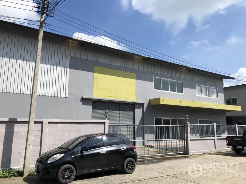 工厂 出租在Pathum Thani - Lat Lum Kaeo两层工厂外立面，灰墙配黄色雨棚，金属大门与停车位。