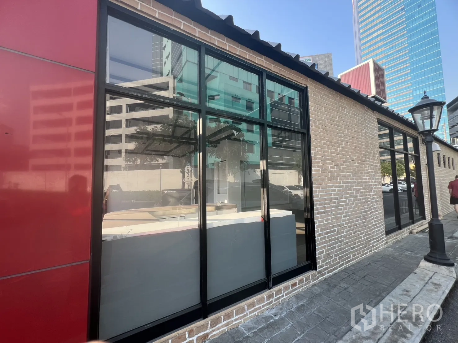 พื้นที่ขายปลีก ให้เช่า - ด้านข้างอาคารผนังก่ออิฐโชว์แนว หน้าต่างกรอบดำและแผงสีแดง.