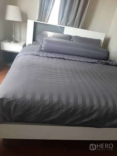Condominium for rent or sale in Huai Khwang, Bangkok - Cozy bedroom with grey bedding, bedside lamp, and curtained window.