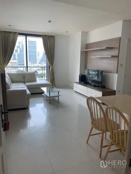 Condominium for rent or sale in Khlong Toei, Bangkok - Living room with L-shaped sofa, TV console and glass coffee table facing a balcony and Bangkok skyline view.