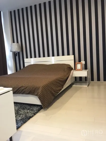 Condominium for rent in Pathum Wan, Bangkok - Primary bedroom featuring a striped accent wall, white bed frame, brown duvet, and bedside table.