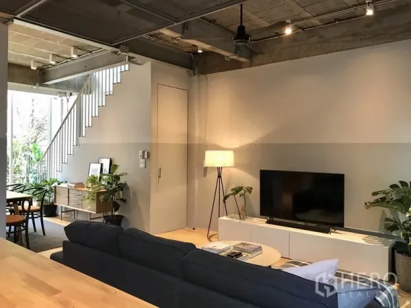 Townhouse for rent in Khlong Toei, Bangkok - TV lounge with sofa, floor lamp and open staircase beneath an exposed ceiling.