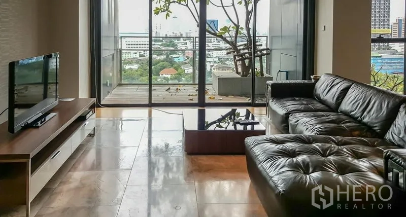 Condominium for rent or sale in Sathon, Bangkok - Bright living room with a leather sectional, TV console, and balcony city view through large sliding glass doors.