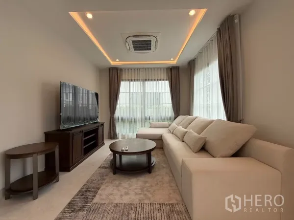 独立式住宅 出租或出售在Bang Phli, Samut Prakan - 明亮客厅，米色转角沙发、电视柜、吊顶氛围灯与纱帘。