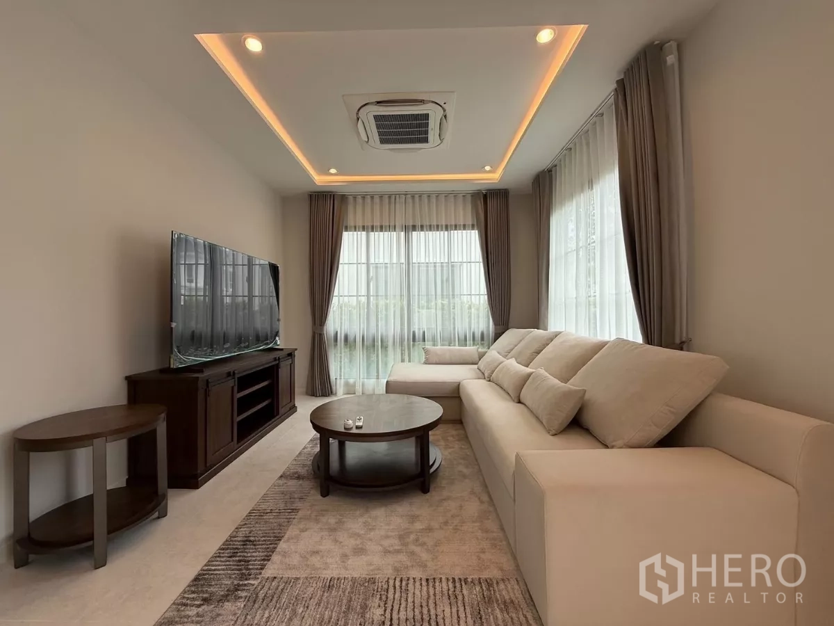 独立式住宅 出租或出售在Bang Phli, Samut Prakan - 明亮客厅，米色转角沙发、电视柜、吊顶氛围灯与纱帘。
