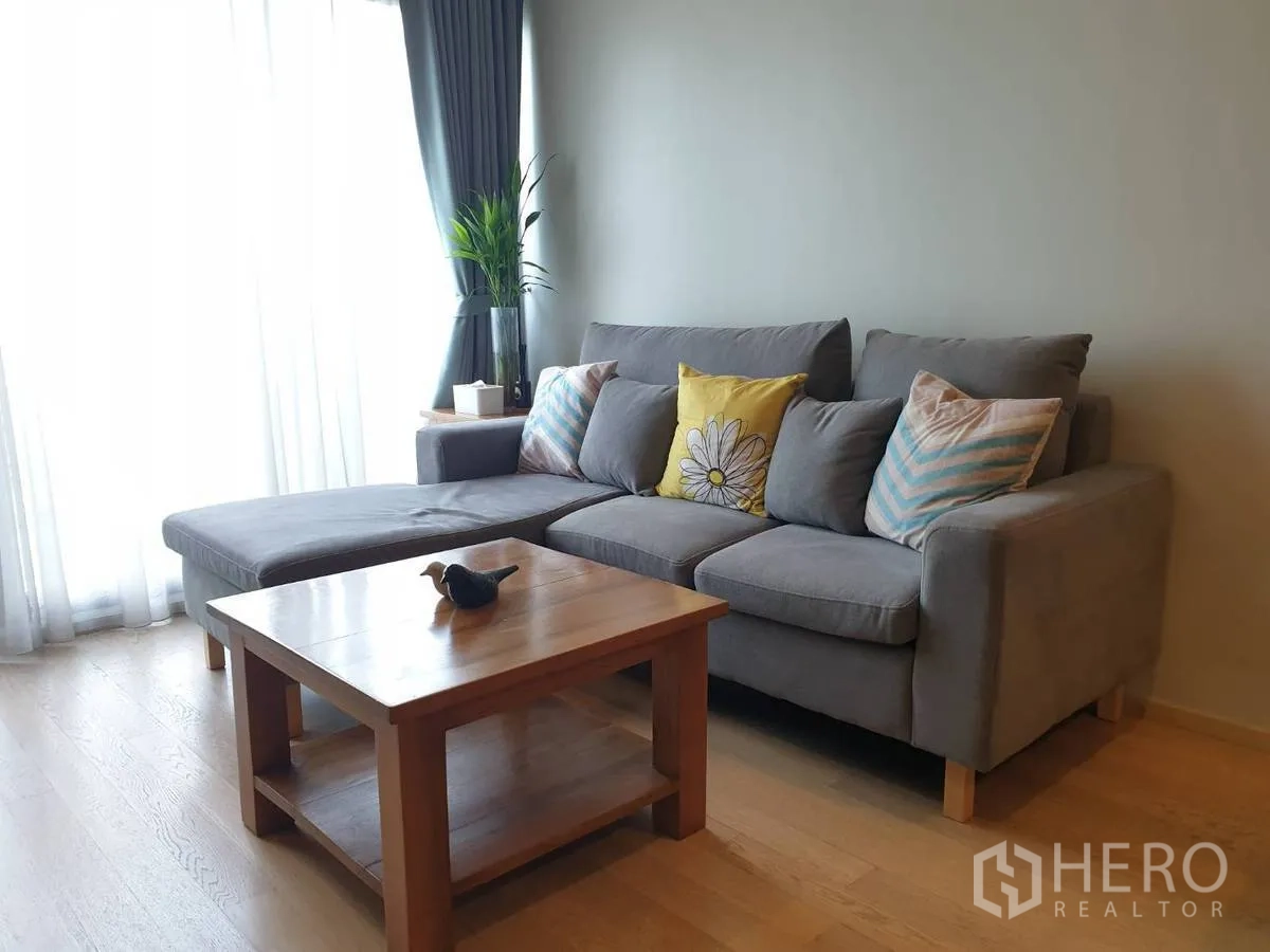 คอนโด ให้เช่าหรือขายในBangkok - โซฟาเข้ามุมข้างหน้าต่างผ้าม่านบานใหญ่ พร้อมโต๊ะกลางไม้และไม้ประดับ