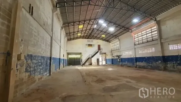 Warehouse for rent in Phra Samut Chedi, Samut Prakan - Deep warehouse bay with high ceiling, a loading door, and a metal staircase leading to a mezzanine office.